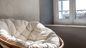 Ein Korbstuhl mit gepolstertem Sitz und Rücken, platziert in der Nähe eines Fensters mit Blick auf einen Balkon.