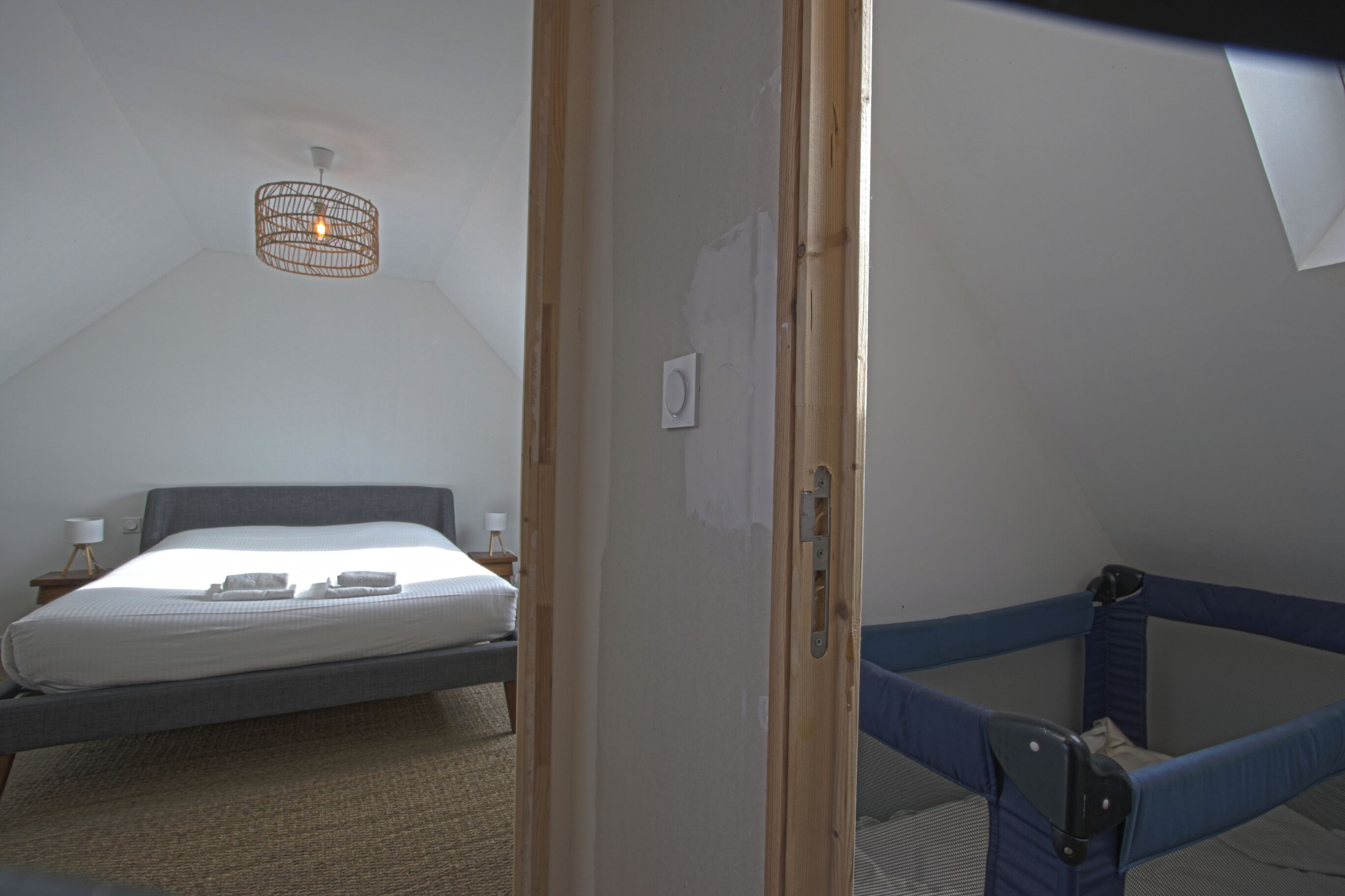 Una habitación con cama, una mesita de noche con lámpara, un aplique en el techo y una puerta que da a una zona con literas.