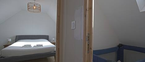 Una habitación con cama, una mesita de noche con lámpara, un aplique en el techo y una puerta que da a una zona con literas.