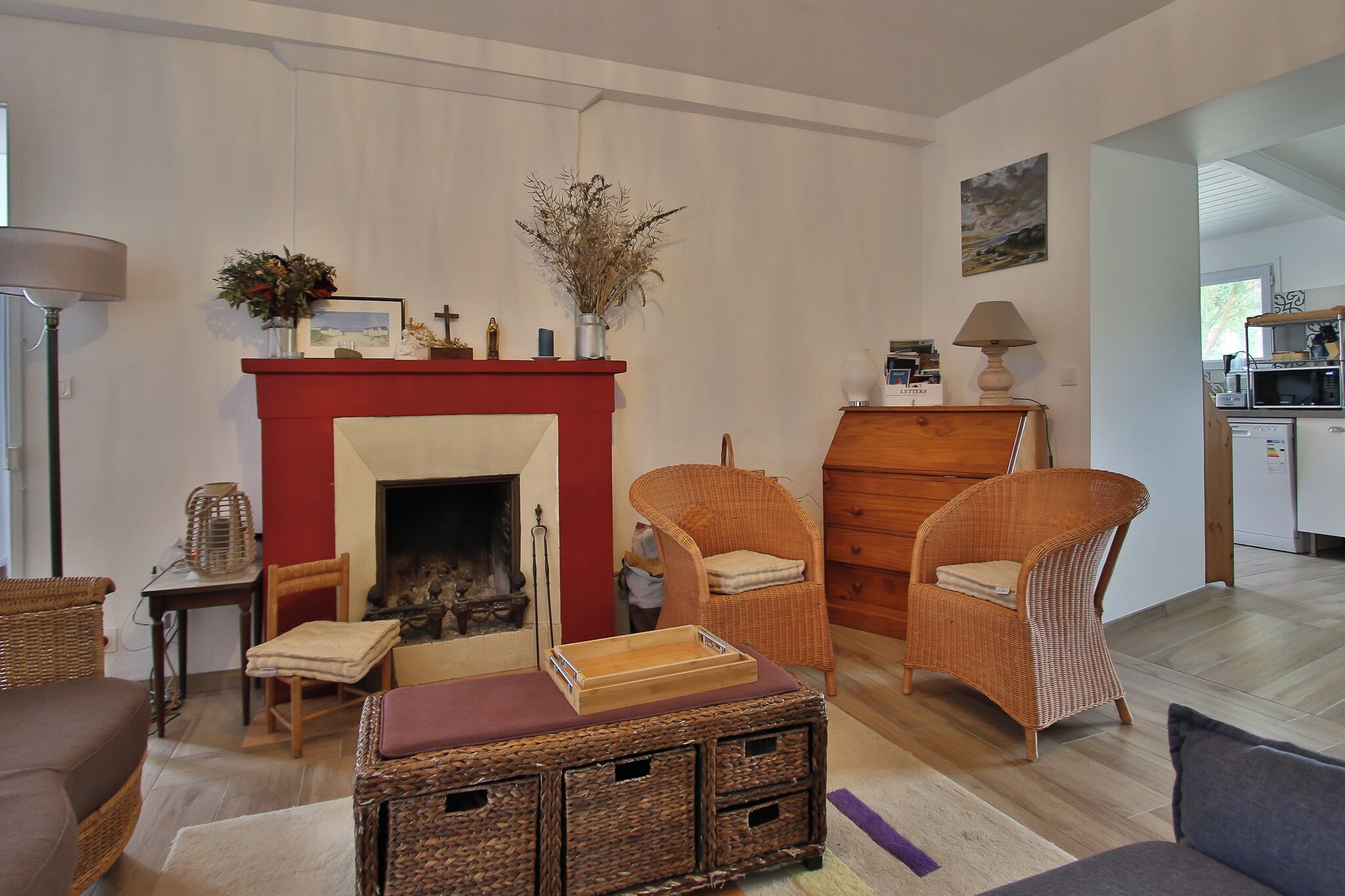 Un salon avec une cheminée, des meubles en osier et une cuisine à l’arrière-plan.