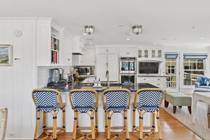 A modern kitchen with white cabinetry, a central island with bar stools, and built-in appliances. - Coastal Luxe at Harris Lane (Kennebunk)