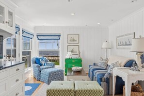 A room with a large window, white walls, and blue patterned furniture. There is a green chest of drawers and a white table with a lamp. - Coastal Luxe at Harris Lane (Kennebunk)