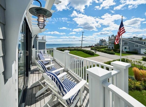 A balcony with white railings and blue-striped chairs, overlooking a residential area with houses and a flag. - Coastal Luxe at Harris Lane (Kennebunk)