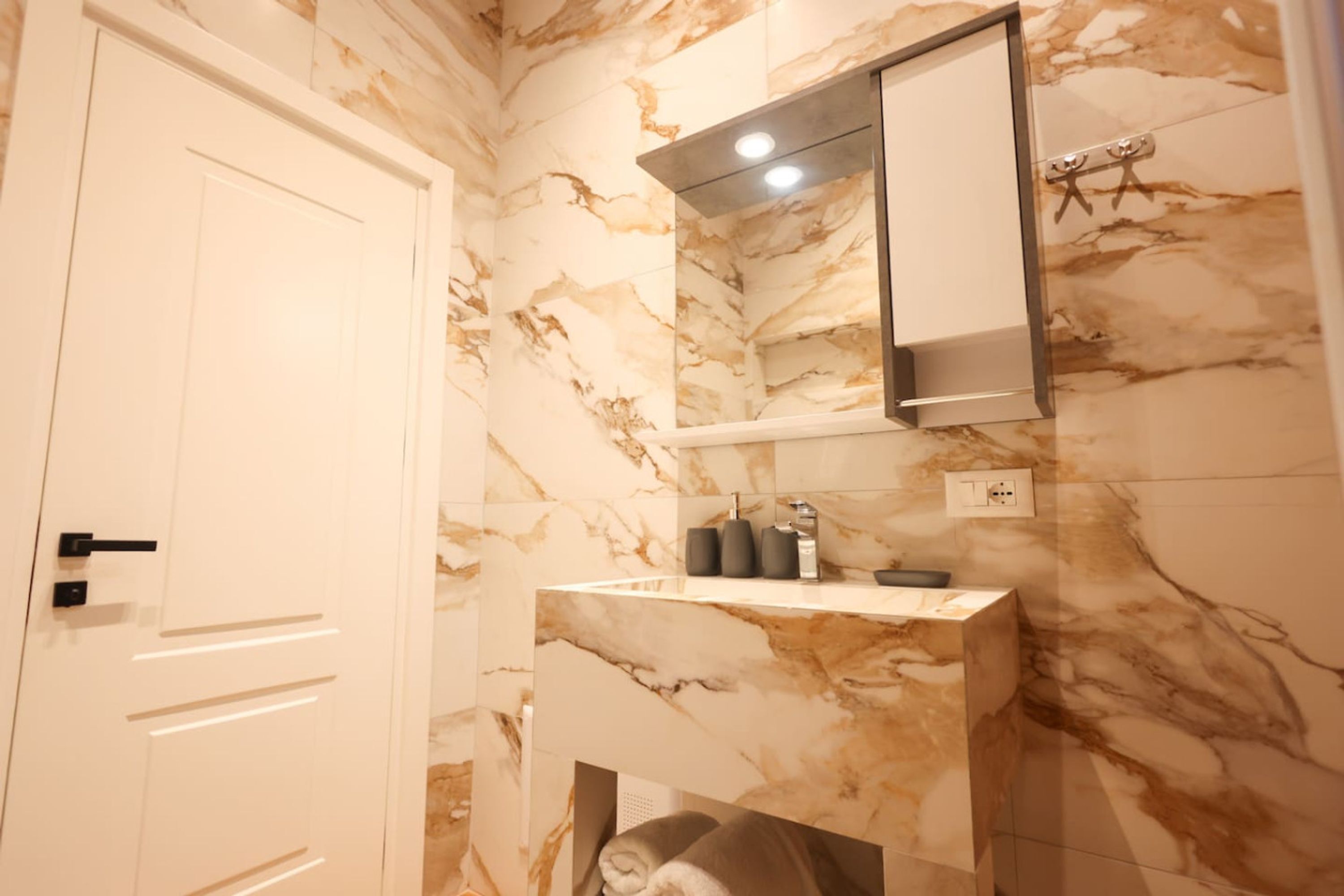 Une salle de bain aux murs de marbre, avec une porte blanche, un miroir et une étagère contenant des articles de toilette.