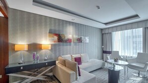 A modern hotel room with a sofa, a dining table, and a coffee table. There are two lamps on a console table, and a large abstract painting on the wall. - Majestic Premier Hotel Bur Dubai (Dubai)