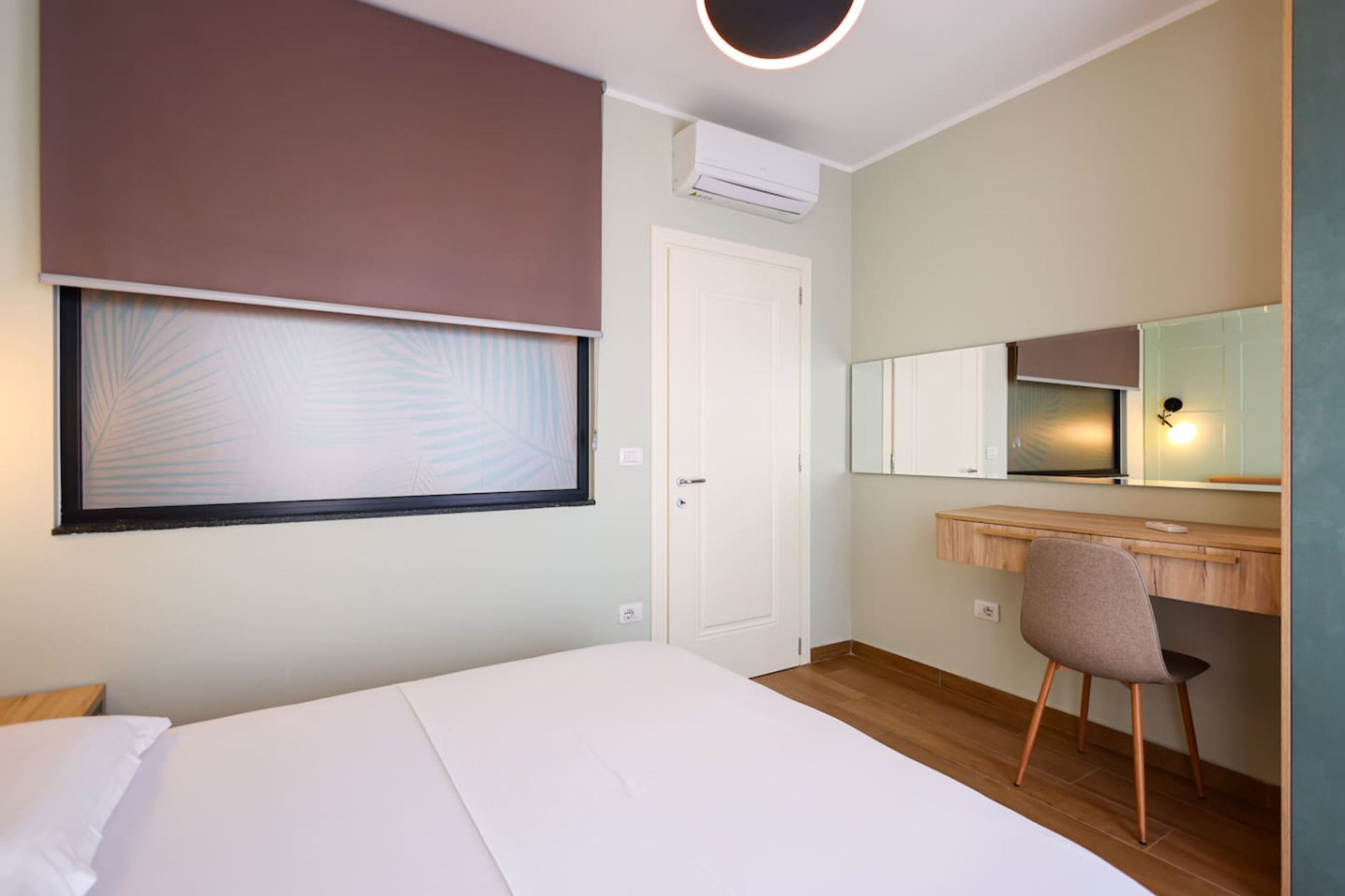 A bedroom with a bed, a desk, a chair, a window with blinds, a door, and a wall-mounted air conditioner.