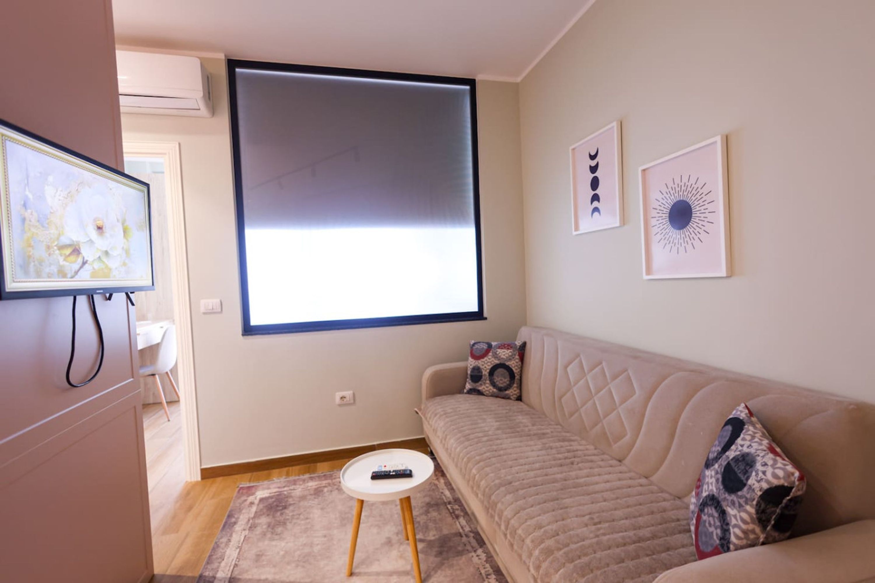 A living room with a beige sofa, a small round coffee table, and a remote control on the table. There are two framed artworks on the wall and a window with blinds.