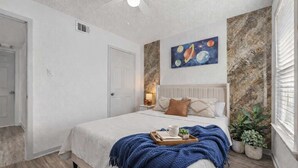 Habitació amb un llit, una tauleta de nit, un quadre a la paret, un ventilador de sostre i una finestra amb persianes.