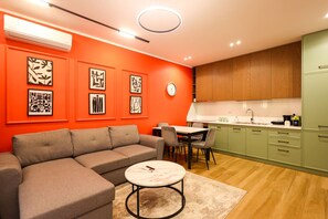 A modern living room with a grey sofa, a round coffee table, and a dining area with a table and chairs. The kitchen features green cabinets and a wooden countertop. - Grandstay Urban One in Durrës (Durrës)