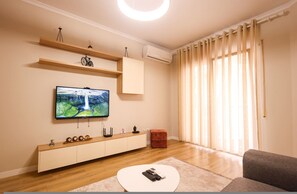 A modern living room with a flat-screen TV, wooden shelves, a red ottoman, and a grey sofa. - Grandstay Urban Five (Durrës)