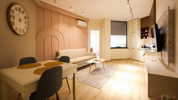 Una sala de estar moderna con sofá, mesa de comedor y sillas. Hay un reloj en la pared y un televisor montado allí.