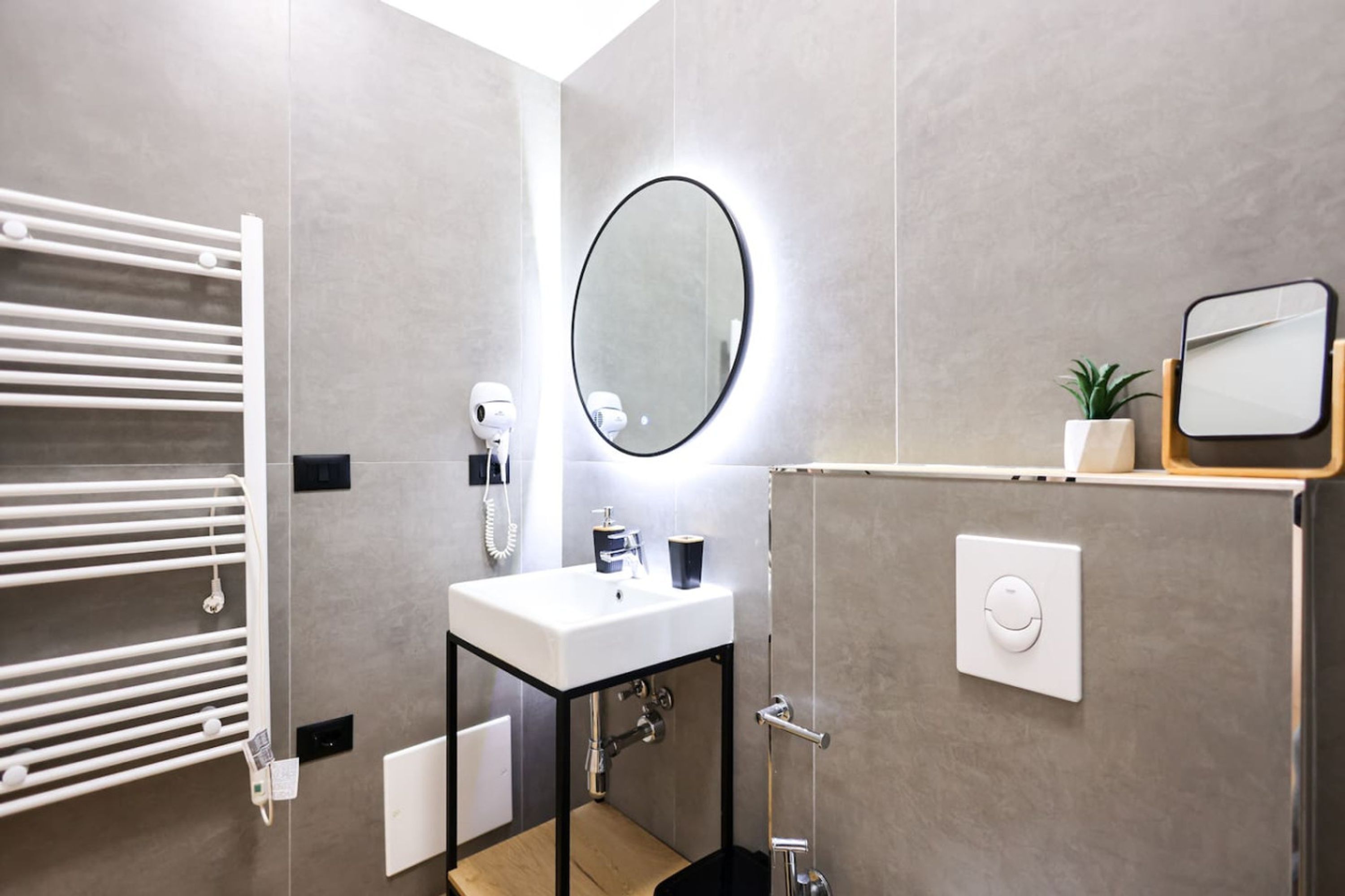 A modern bathroom with a white sink, a round mirror with a light, a towel rack, a handheld showerhead, and a small plant on a shelf.