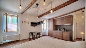 Un dormitorio moderno con una cama grande, una mesita pequeña y sillas, una zona de cocina con armarios de madera, y un televisor fijado a la pared.