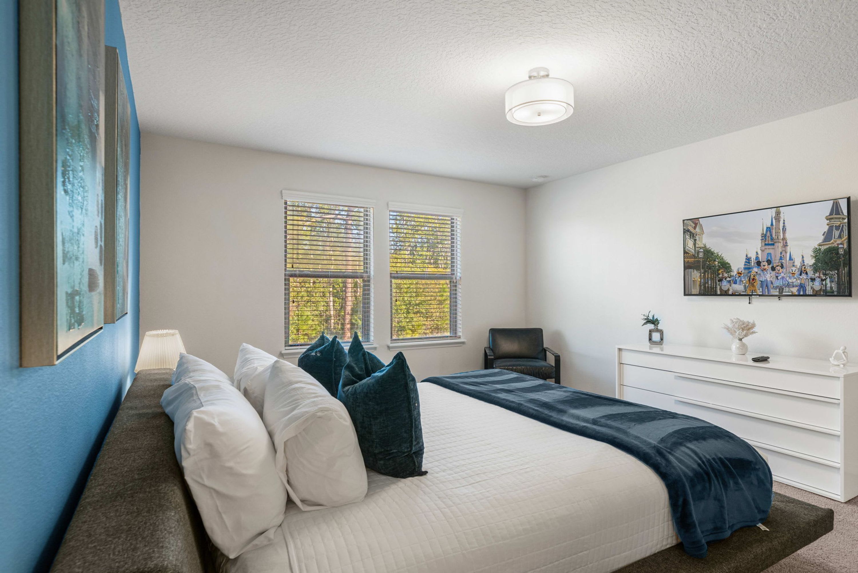 A bedroom with a bed, a desk, and a chair. There are windows with blinds, a painting on the wall, and a ceiling light.