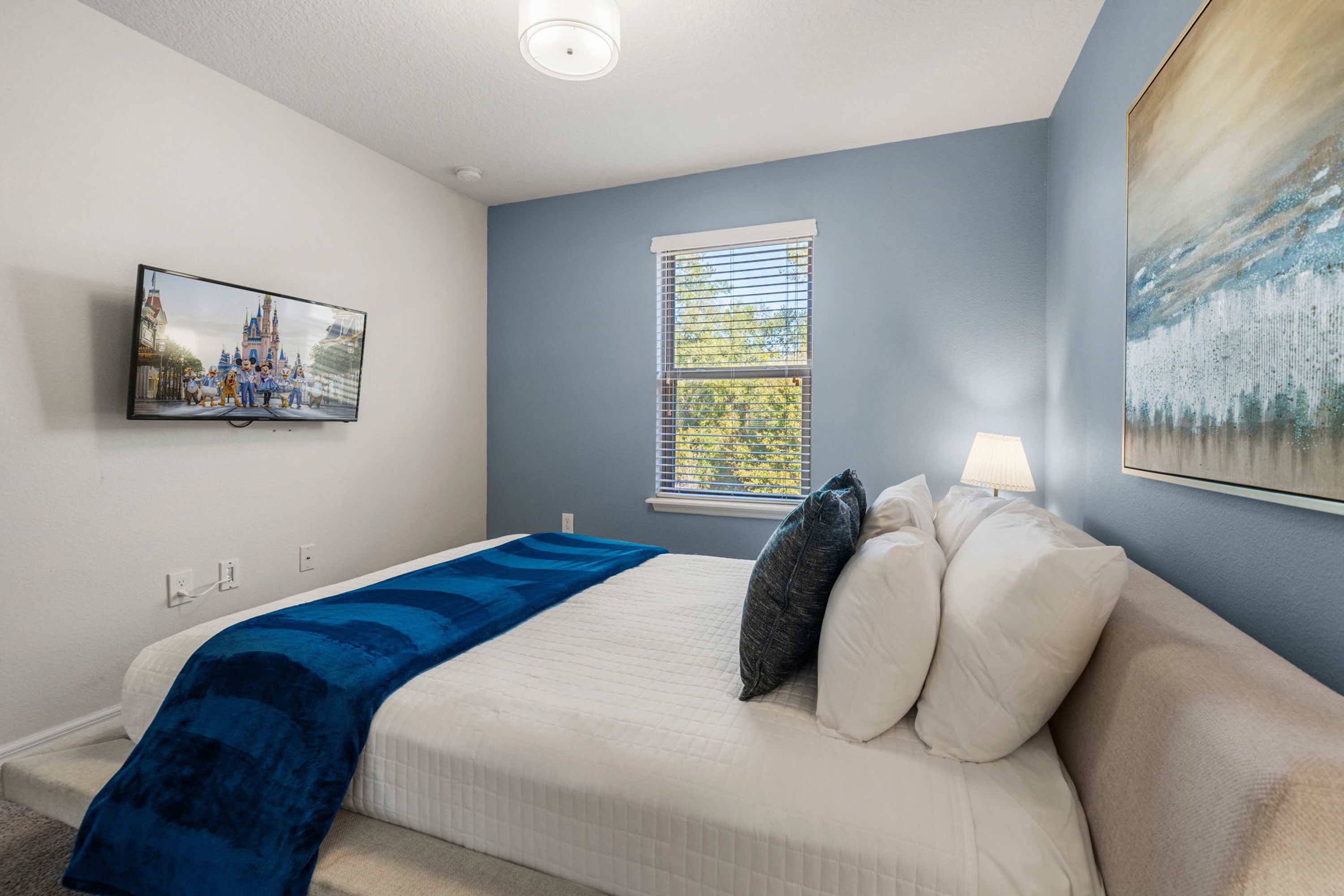 A bedroom with a bed, a TV mounted on the wall, a window with blinds, and a painting on the wall.