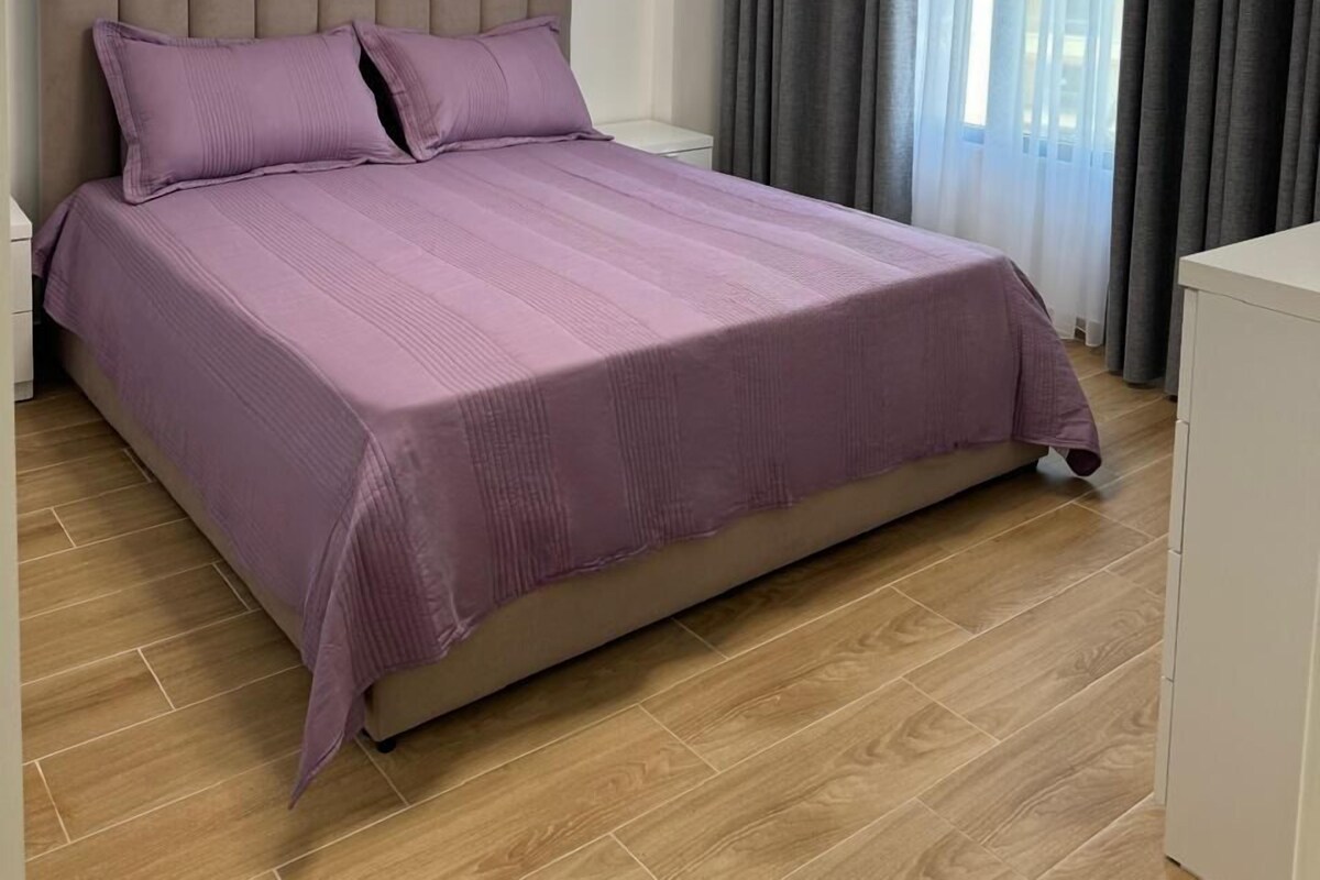 A modern bedroom with a large bed, a bedside table, a hanging light fixture, and a window with curtains.