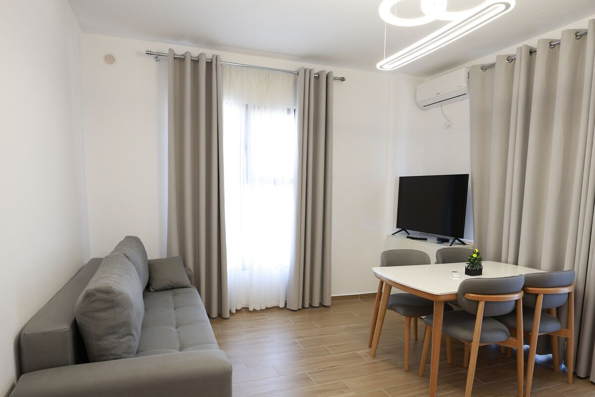 A modern living room with a sofa, a dining table with chairs, a flat-screen TV, and a ceiling light fixture.