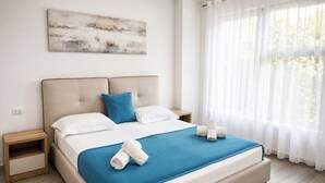 A modern bedroom with a large bed, two bedside tables, a blue bedspread, white pillows, and a view of trees through sheer curtains. - Stay By Ilvesta Potam (Himarë)