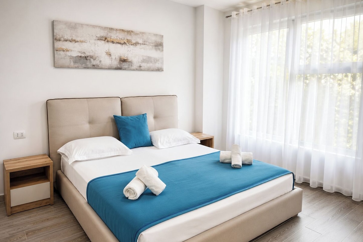 A modern bedroom with a large bed, two bedside tables, a blue bedspread, white pillows, and a view of trees through sheer curtains.
