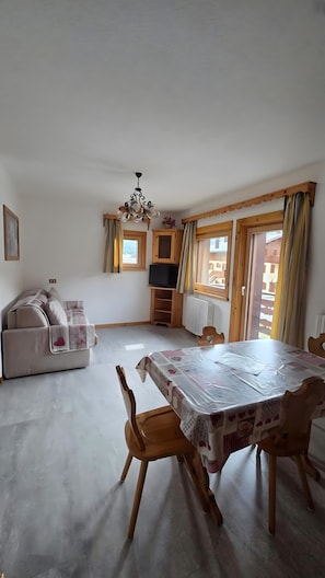 Ein Zimmer mit Esstisch, Stühlen, einer Couch, einem Fernseher und einem Fenster mit Vorhängen.