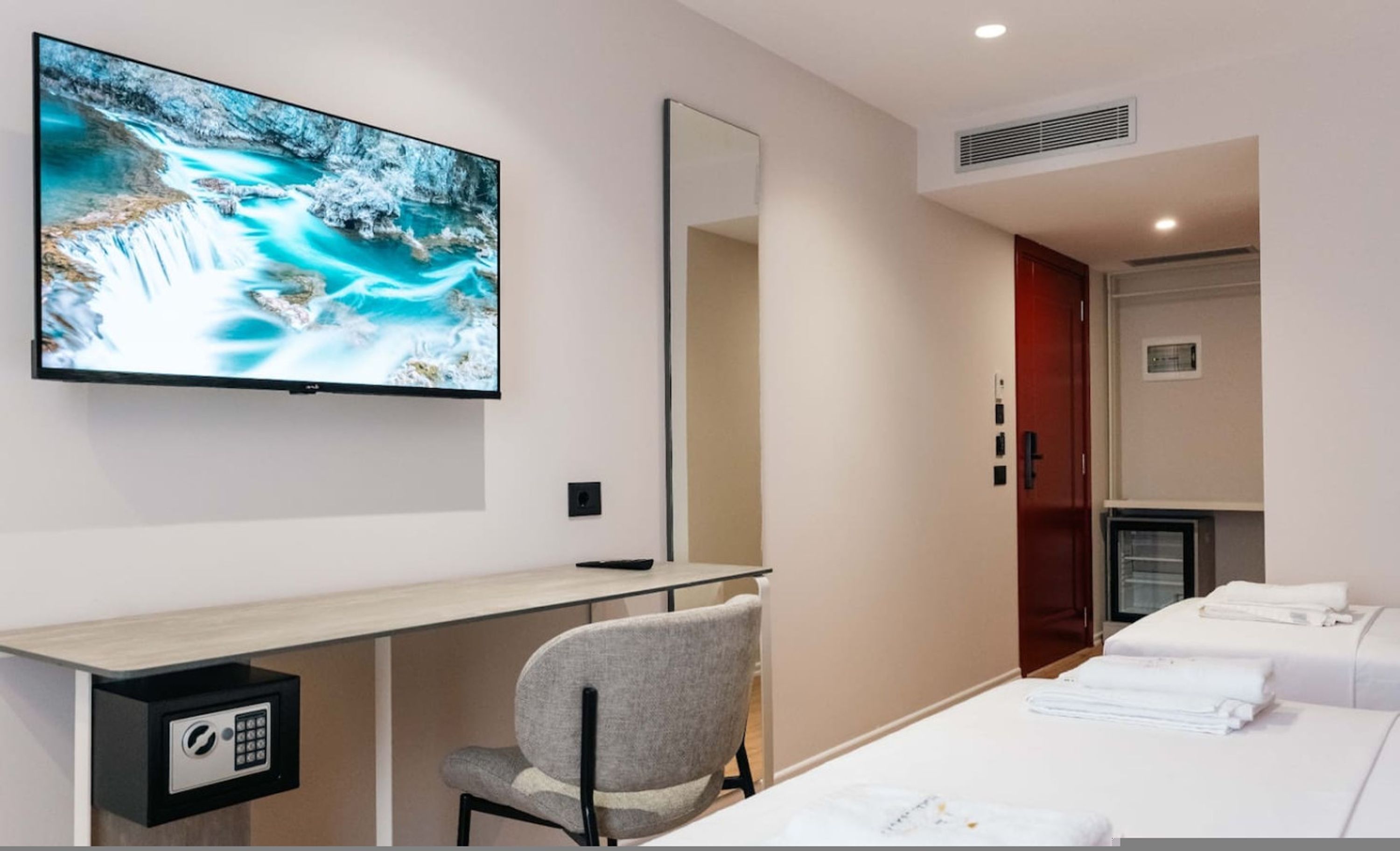 A hotel room with a large TV on the wall, a desk with a chair, a bed with towels, and a mirror.