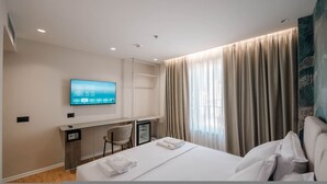 Hotellihuone, jossa on sänky, työpöytä tietokoneella, seinälle asennettu televisio ja suuri ikkuna, jossa on verhot.