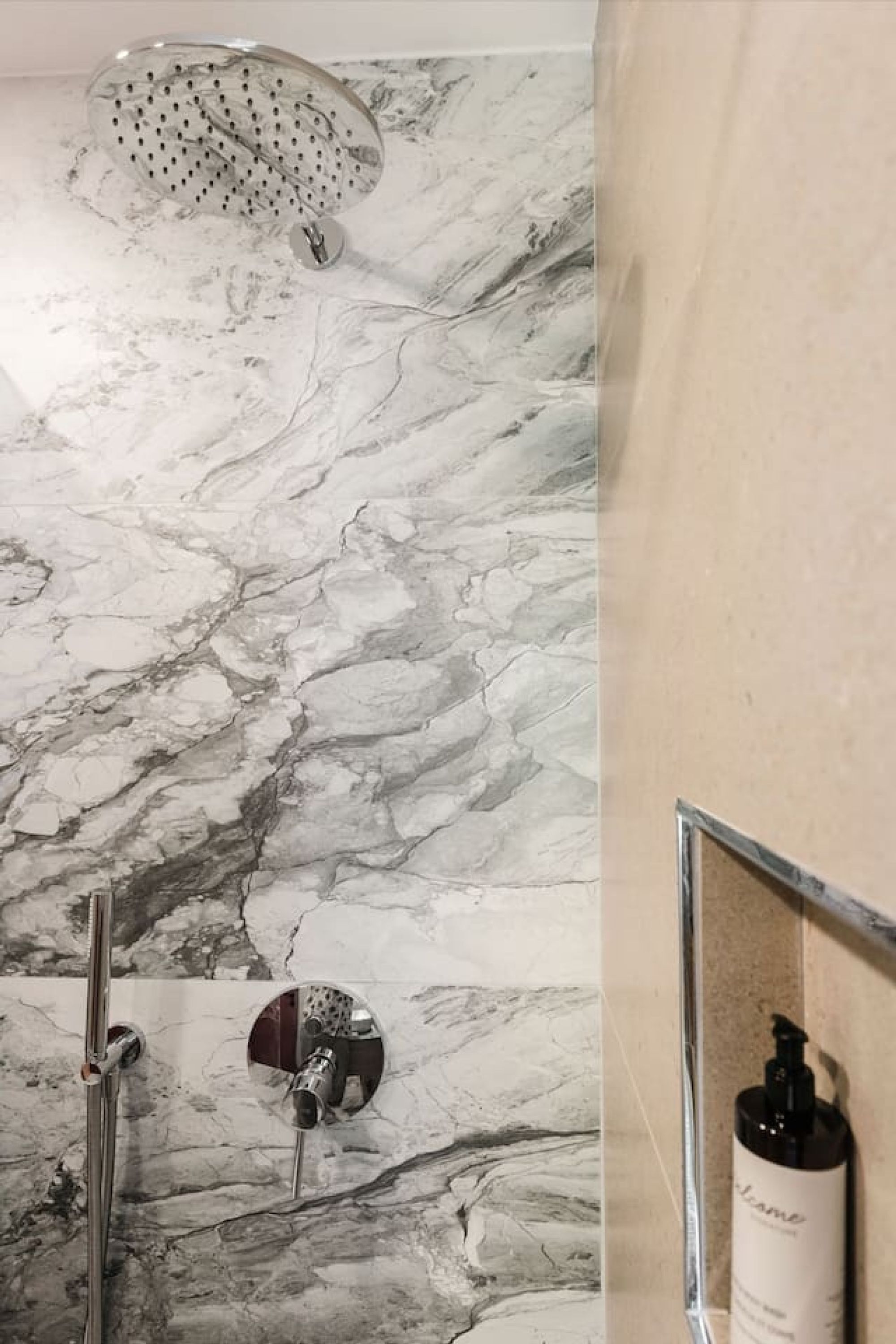 A bathroom with a marble wall, a showerhead, a faucet, and a framed bottle of shampoo.