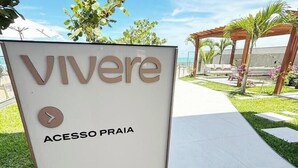 Opastekyltti, jossa lukee ”Vivere Acesso Praia”, ja sen vierestä kulkee polku, joka johtaa katettuun ulko-oleskelualueeseen.