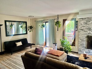 Un salon avec un canapé, une table basse et une cheminée. On y trouve des plantes, des rideaux et un miroir accroché aux murs.