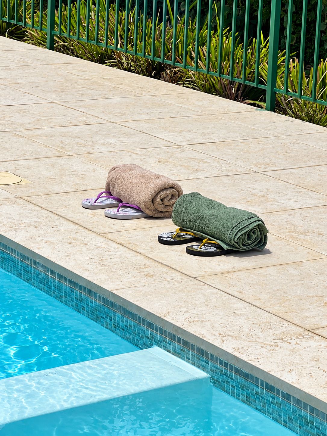 A poolside area with a tiled deck, a pool, and a green metal fence.
