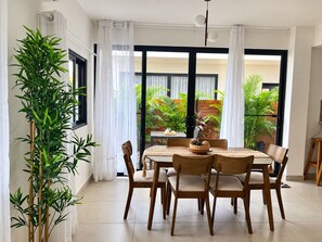 Ein Essbereich mit Tisch und Stühlen, einer großen Kübelpflanze und Blick auf einen Garten durch eine Glastür.