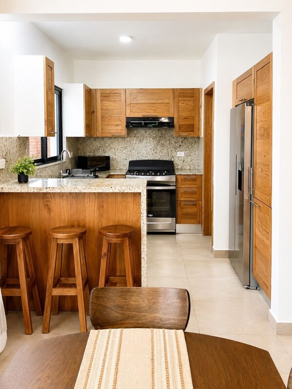 A modern kitchen with wooden cabinets, a stainless steel refrigerator, a stove, and bar stools.
