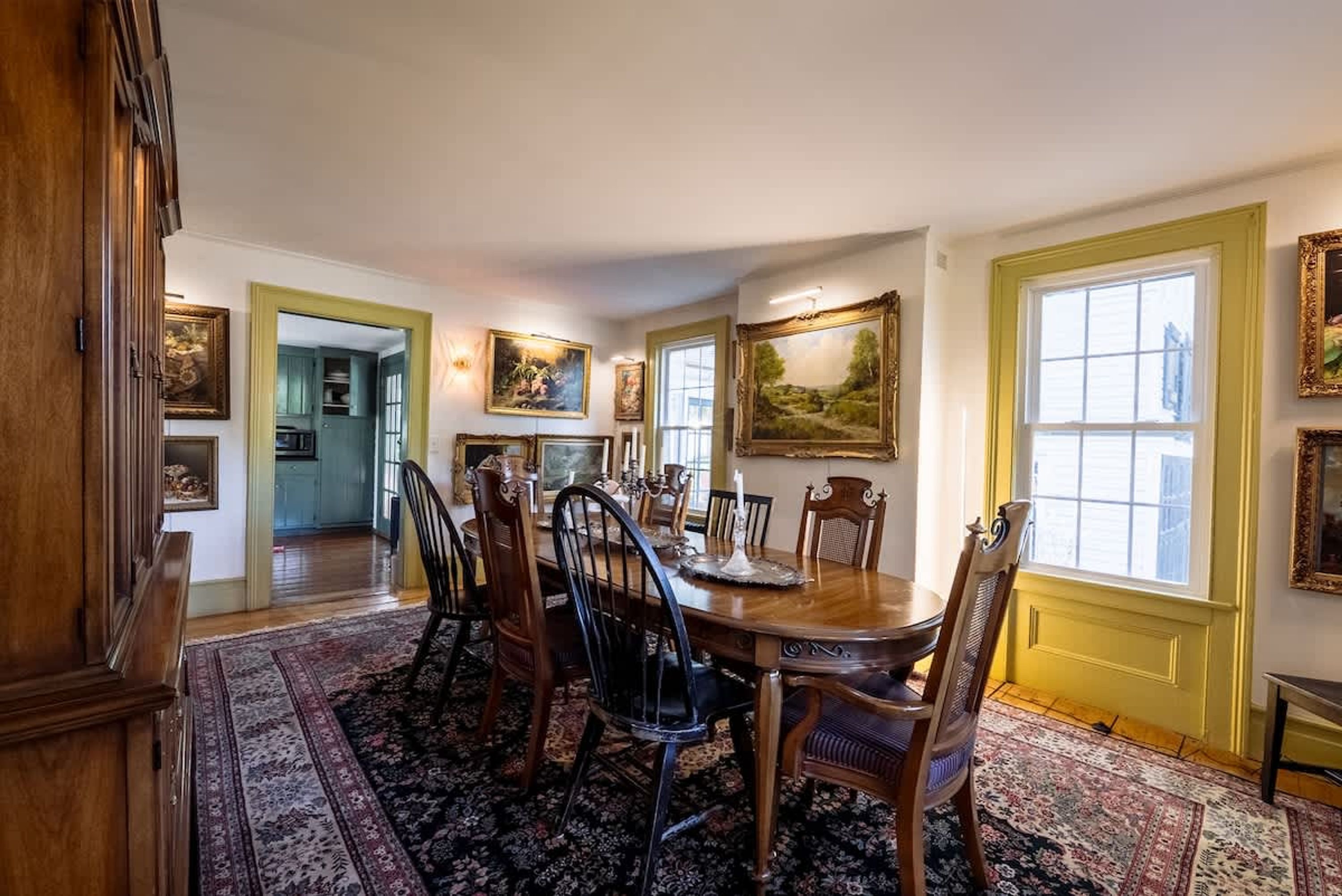 Une salle à manger avec une table en bois, des chaises et un tapis. Les murs sont ornés de tableaux et il y a une fenêtre avec un cadre jaune.