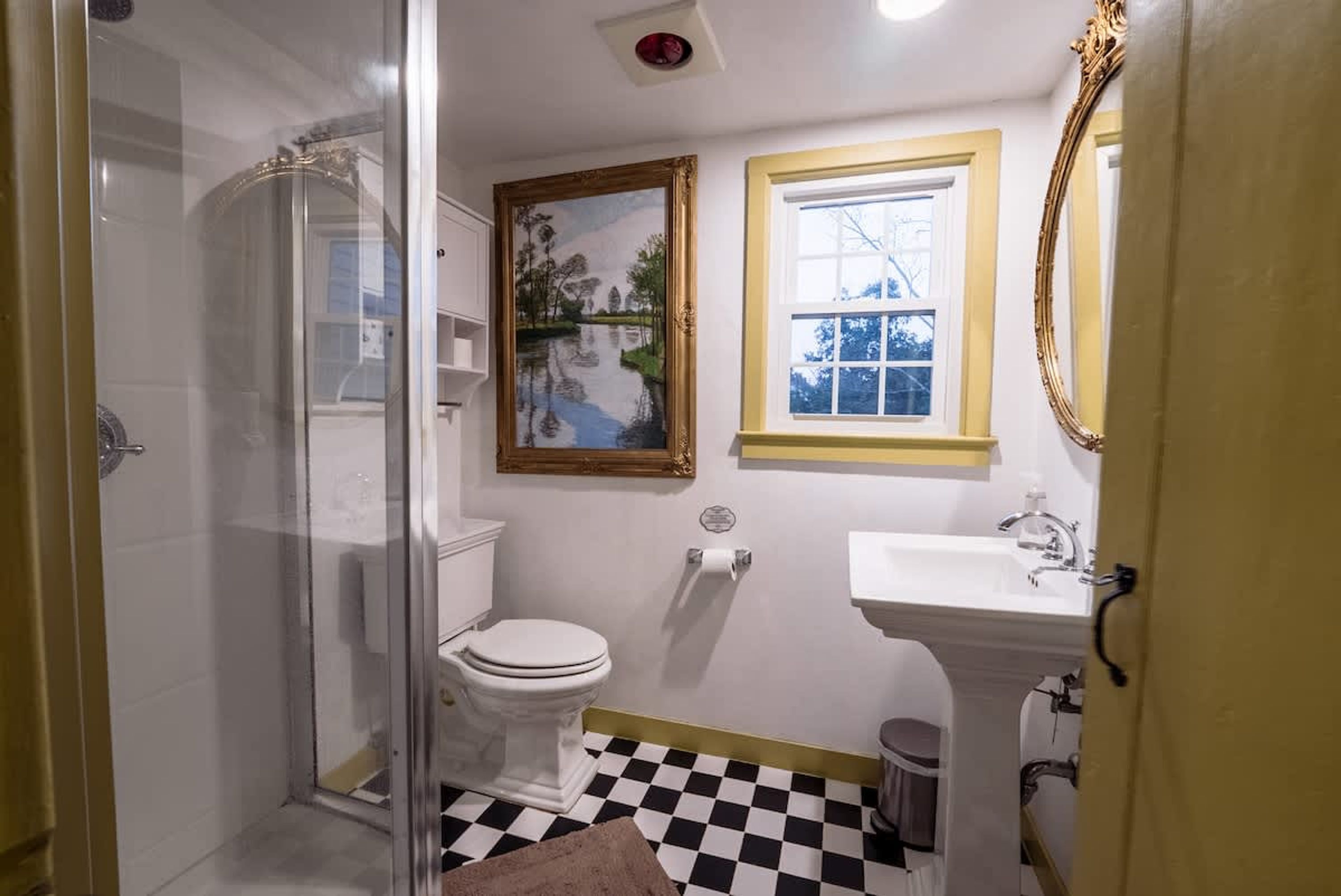 A bathroom with a glass shower enclosure, a toilet, a sink, a framed painting, and a window with a view of trees.