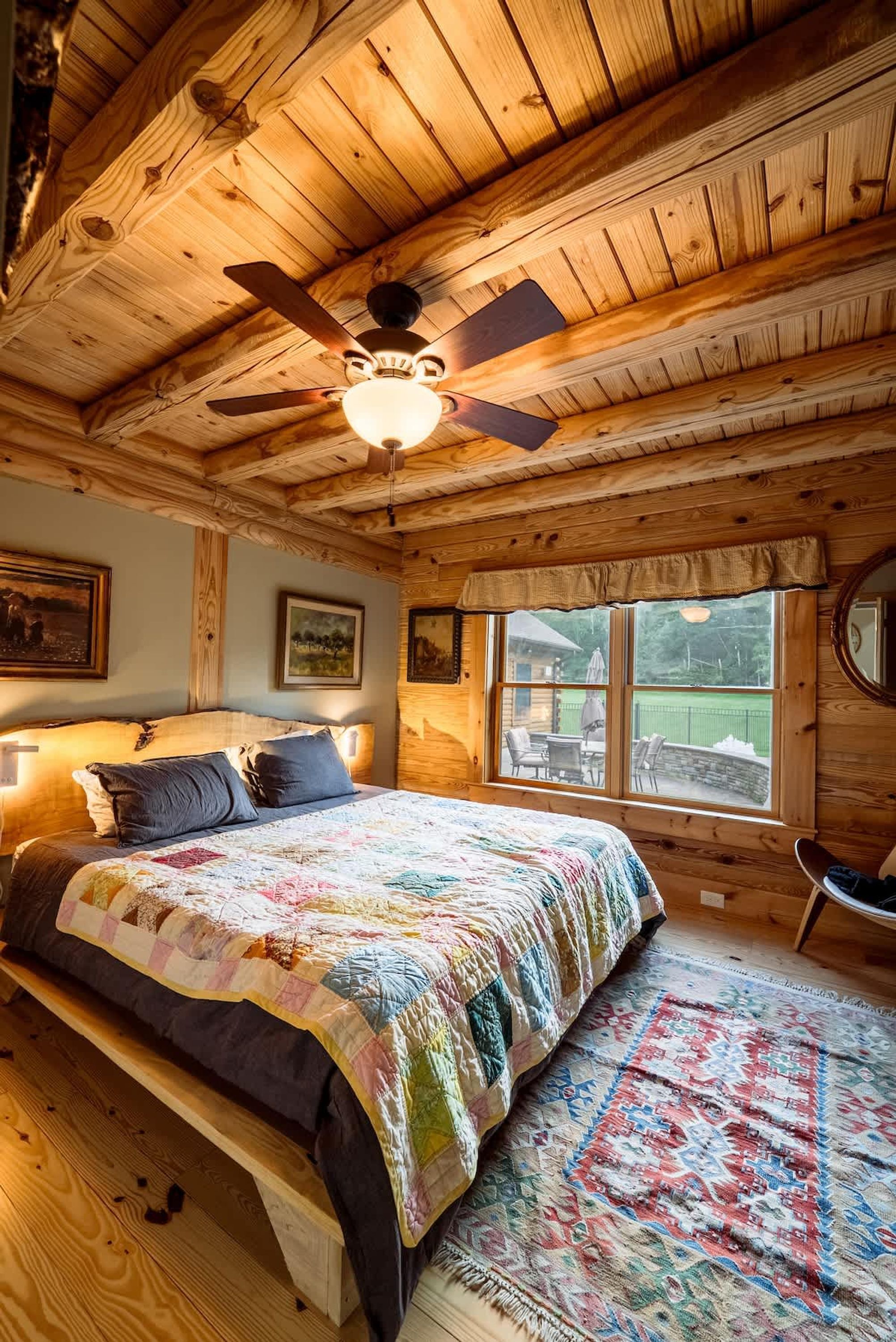 A wooden cabin bedroom with a large bed, a ceiling fan, and a window overlooking a deck.