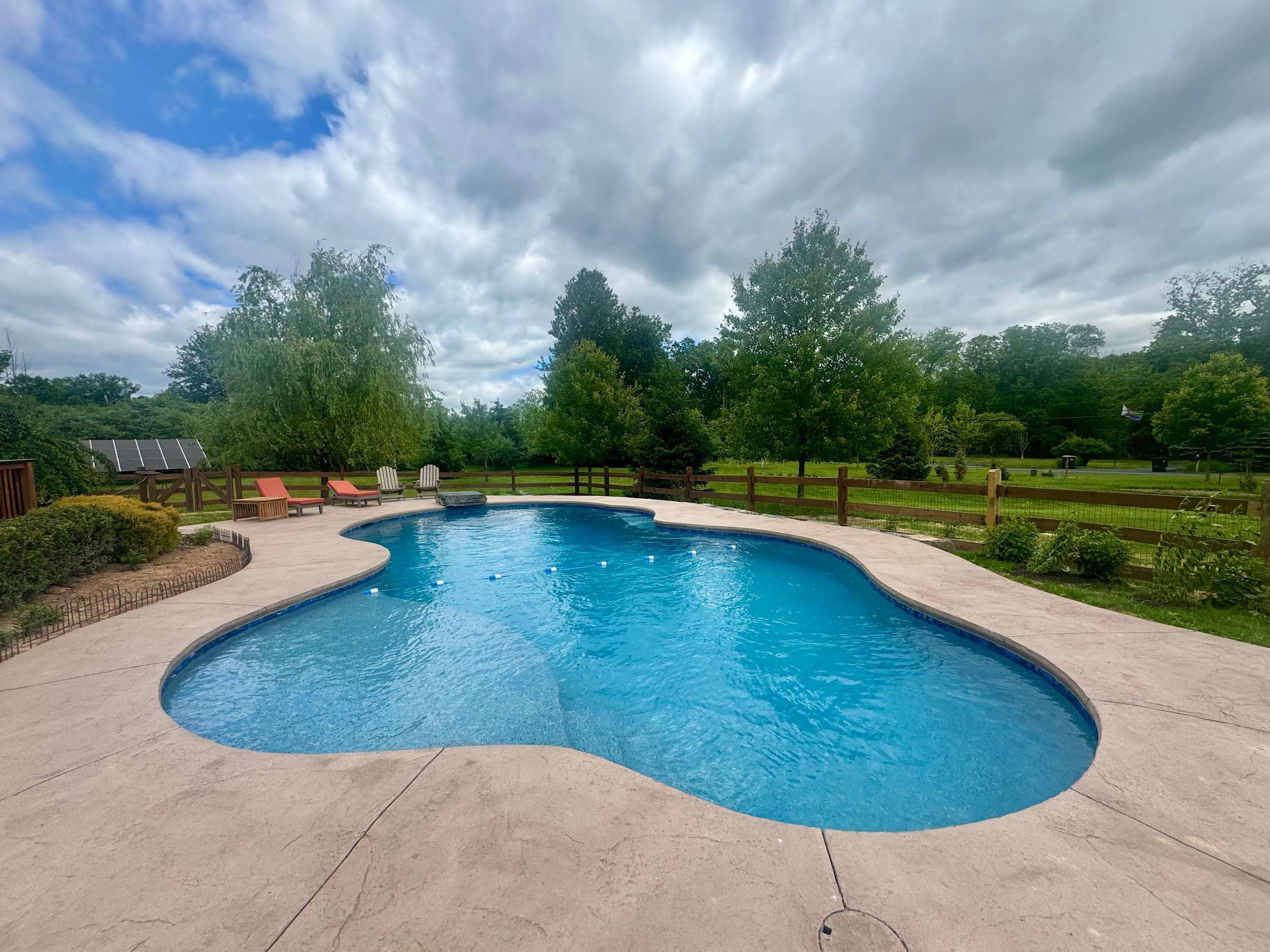 A swimming pool with a curved edge, surrounded by a wooden fence, and a grassy area with trees in the background.