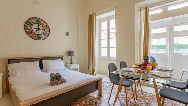 Een hotelkamer met een bed, een eettafel met stoelen, een klok aan de muur en een raam met gordijnen.
