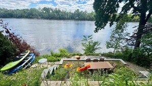 A lakeside patio with a table and chairs, surrounded by greenery and a view of the lake. - Charming   Whimsical Historic River Home (Hopewell Township)