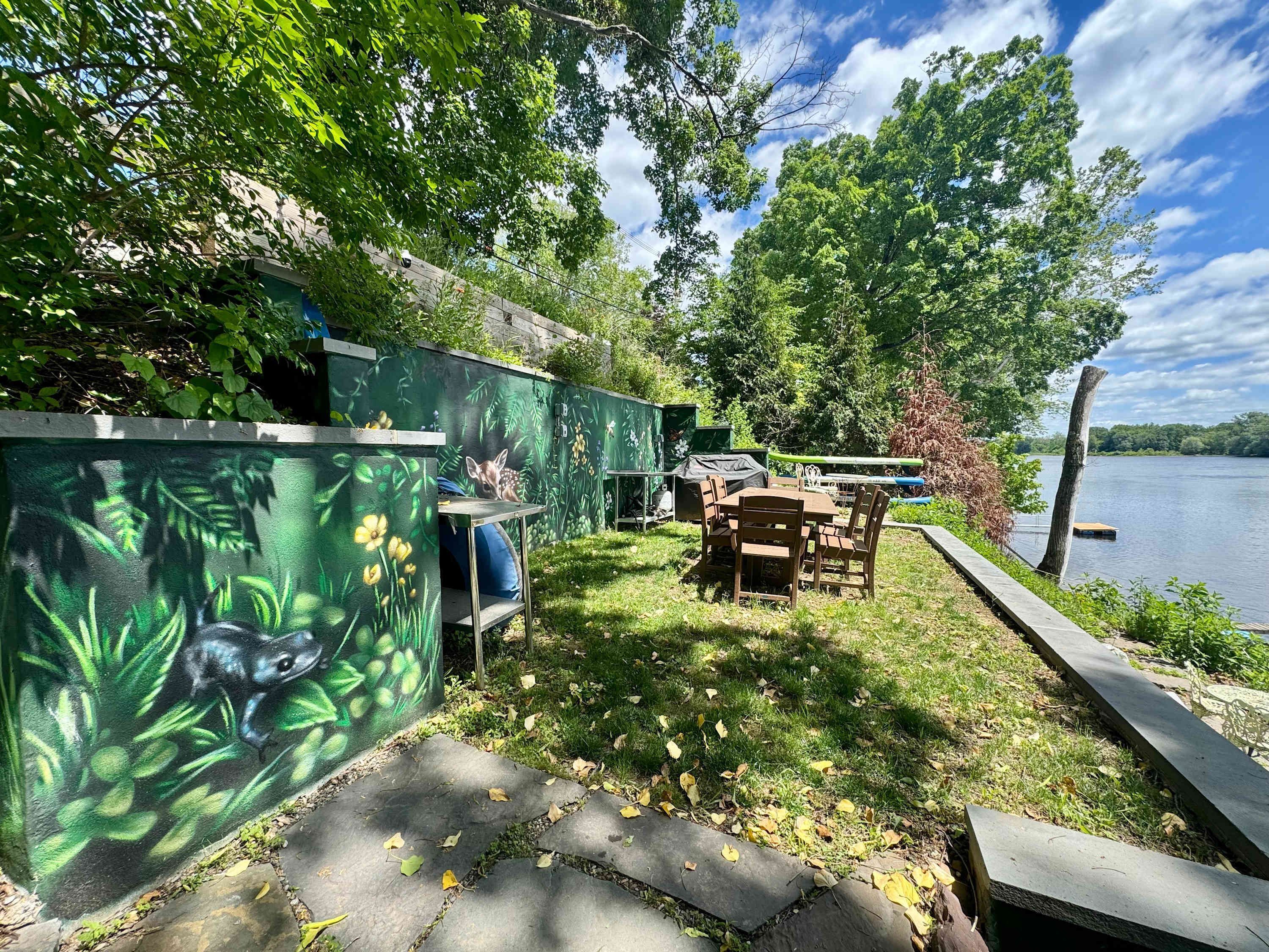 A lakeside patio with a table and chairs, a painted wall with nature scenes, and a dock extending into the water.