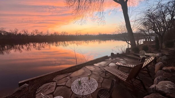 A lakeside patio with stone flooring, outdoor seating, and a view of the sunset over the water. - Charming   Whimsical Historic River Home (Hopewell Township)