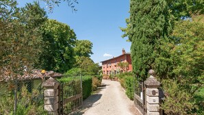 En grussti fører hen til en rød bygning med mange vinduer, omgivet af grønt og stenporte.