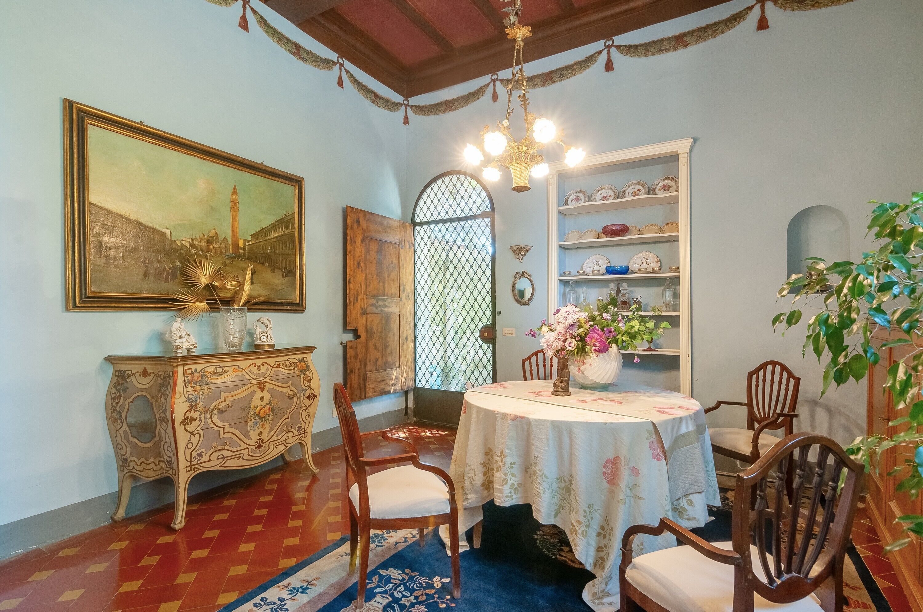 A dining area with a round table, chairs, a chandelier, a cabinet with decorative items, a painting, and a window with a lattice design.
