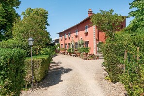 A red building with a gravel path, surrounded by greenery and potted plants. - Elegant room in 15th-century villa with private bathroom and pool (Grassina)