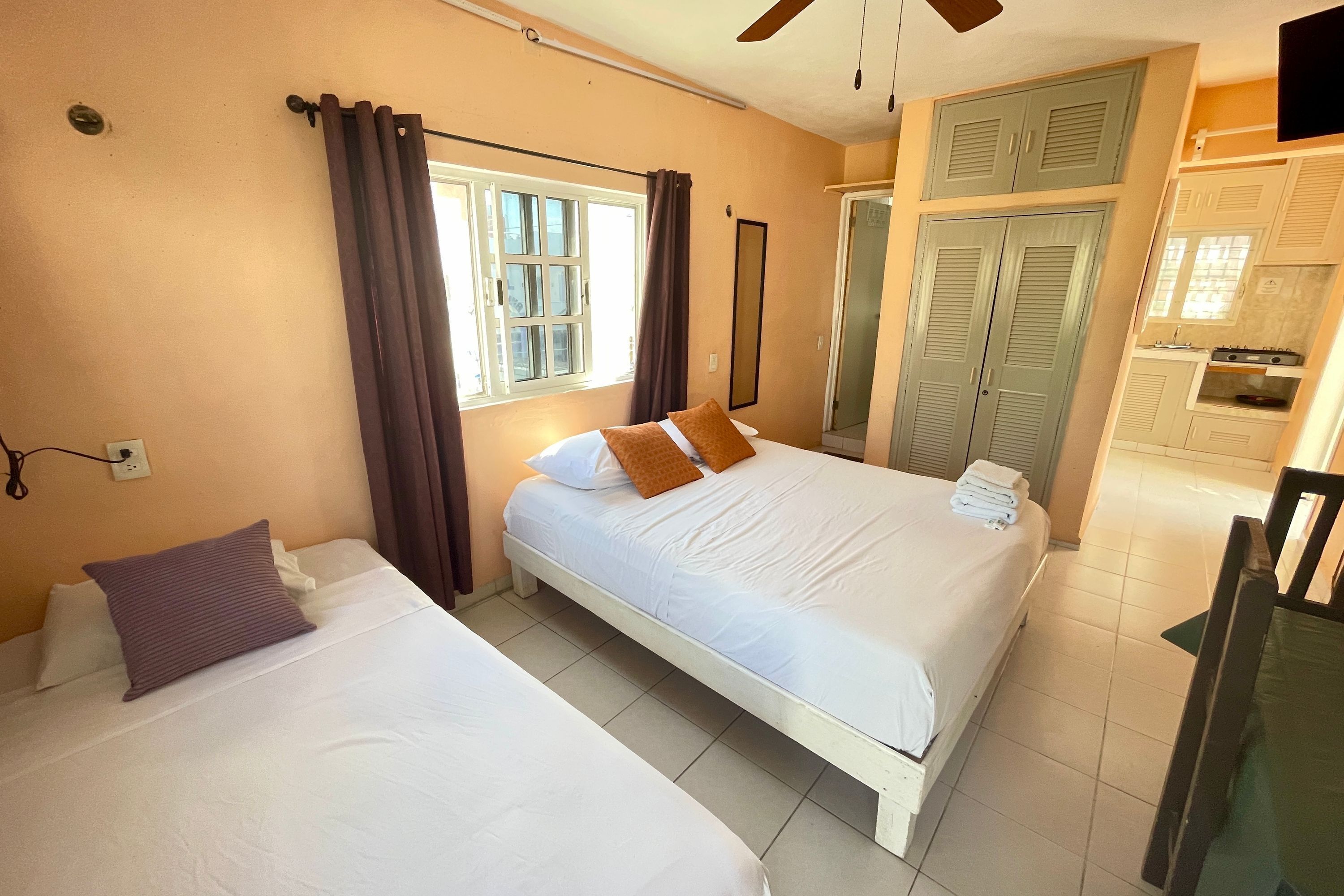 A hotel room with two beds, a window with curtains, a ceiling fan, and a kitchenette.