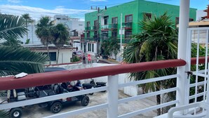 Ein Balkon mit roter Brüstung, der auf eine Straße mit geparkten Fahrzeugen und einem grünen Gebäude im Hintergrund blickt.