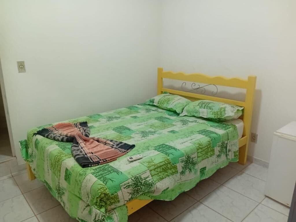 A small, single-bed room with a yellow headboard, green and white patterned bedding, and a white bedside table.