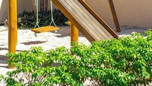 Eine hölzerne Spielanlage mit Rutsche und Schaukeln, umgeben von Grünflächen und Sandboden.