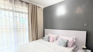 Een slaapkamer met een groot bed, witte beddengoed, een nachtkastje, een wandlamp en dunne gordijnen.