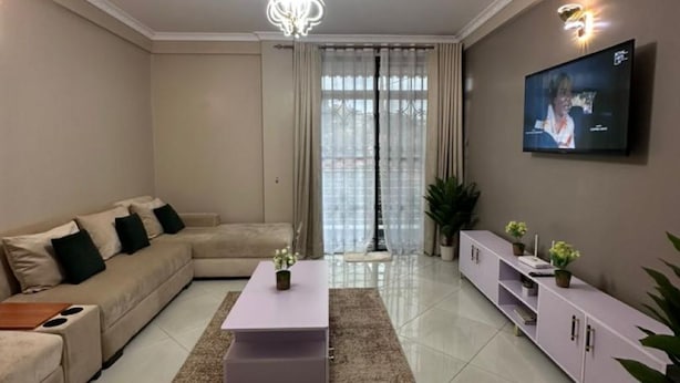 Een moderne woonkamer met een beige bank, een witte salontafel, een aan de muur bevestigde televisie en een kroonluchter.