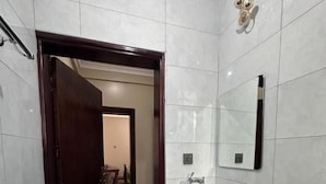 Een moderne badkamer met een witte wastafel, spiegel en toilet. Een houten deur leidt naar een andere kamer, en er liggen een paar slippers op de vloer.